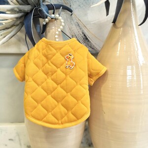 May include: A quilted yellow dog jacket with a diamond pattern and a giraffe applique. The jacket has short sleeves and a rounded neckline. The background includes decorative vases.