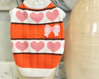 May include: An orange and white dog sweater with pink heart patterns and a pink bow applique. The sweater has a ribbed texture and a white collar and cuffs. A decorative item for pets.