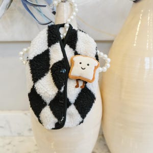 Könnte beinhalten: Eine schwarz-weiß karierte Jacke mit einer kleinen, lächelnden Toast-förmigen Anstecknadel. Die Jacke hat einen Knopfverschluss und ist aus einem weichen, strukturierten Material gefertigt. Eine Perlenkette ist um die Jacke drapiert.