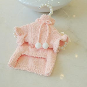 May include: A light pink pet sweater with a hood and two white pom-poms. The sweater has ruffled edges and is hanging from a pearl-beaded hanger. The sweater is made of a soft, fuzzy material.