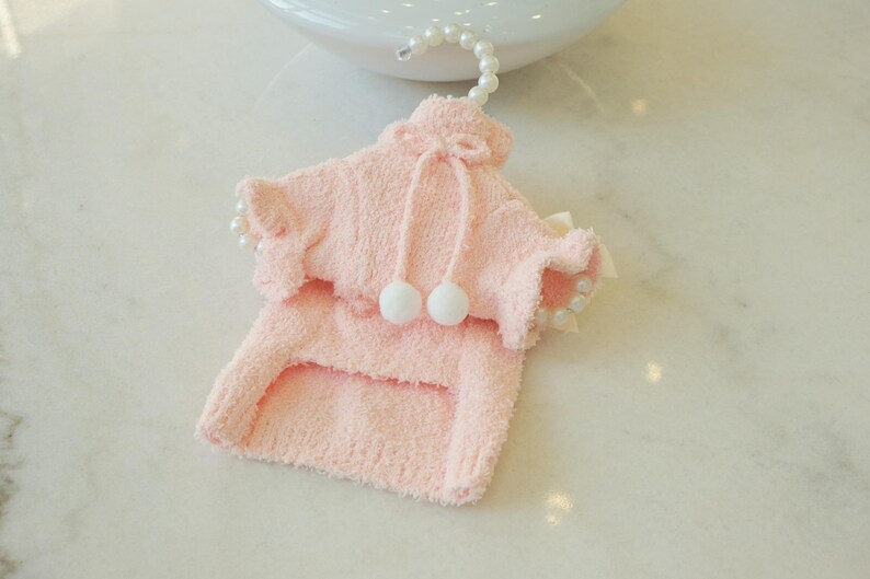 May include: A light pink pet sweater with a hood and two white pom-poms. The sweater has ruffled edges and is hanging from a pearl-beaded hanger. The sweater is made of a soft, fuzzy material.