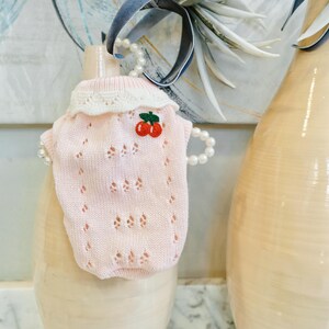 May include: A light pink knitted pet sweater with a white lace collar and cherry applique. The sweater has a delicate openwork design and is displayed on a beige vase. A pearl necklace is draped over the vase.