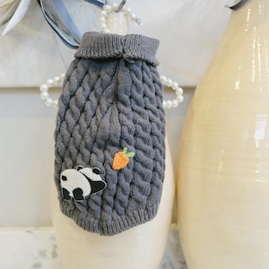 May include: A gray cable-knit dog sweater with a turtleneck collar. The sweater features a panda and carrot applique. The sweater is designed to fit small dogs and is made of a soft, warm material.