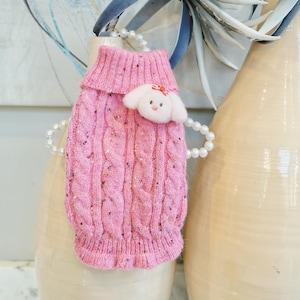 Peut inclure: Un pull rose pour chien en tricot torsadé avec une application de chien en feutre blanc. Le pull a un col roulé et un ourlet à volants. L'application de chien a un nœud rouge. Le pull est décoré de mouchetures de couleur.