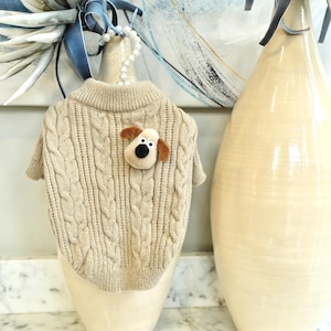 May include: A beige cable-knit dog sweater with a small felt dog applique. The sweater has a round neck and short sleeves. The dog applique is tan and black. The sweater is displayed on a cream-colored vase.