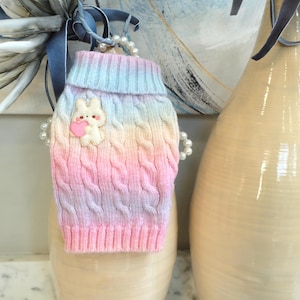 May include: A pastel rainbow-colored dog sweater with a cable knit design. The sweater features a white bunny applique holding a pink heart. The sweater has a turtleneck and is decorated with faux pearls.