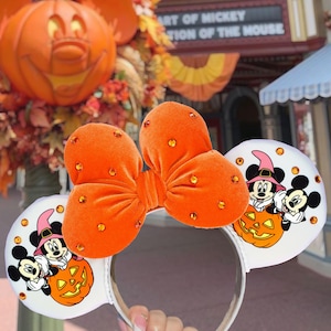 May include: A headband featuring two white ears with a graphic of Mickey and Minnie Mouse in witch hats sitting on a pumpkin. A large orange velvet bow with embedded orange rhinestones sits between the ears. The ears are also accented with orange rhinestones.