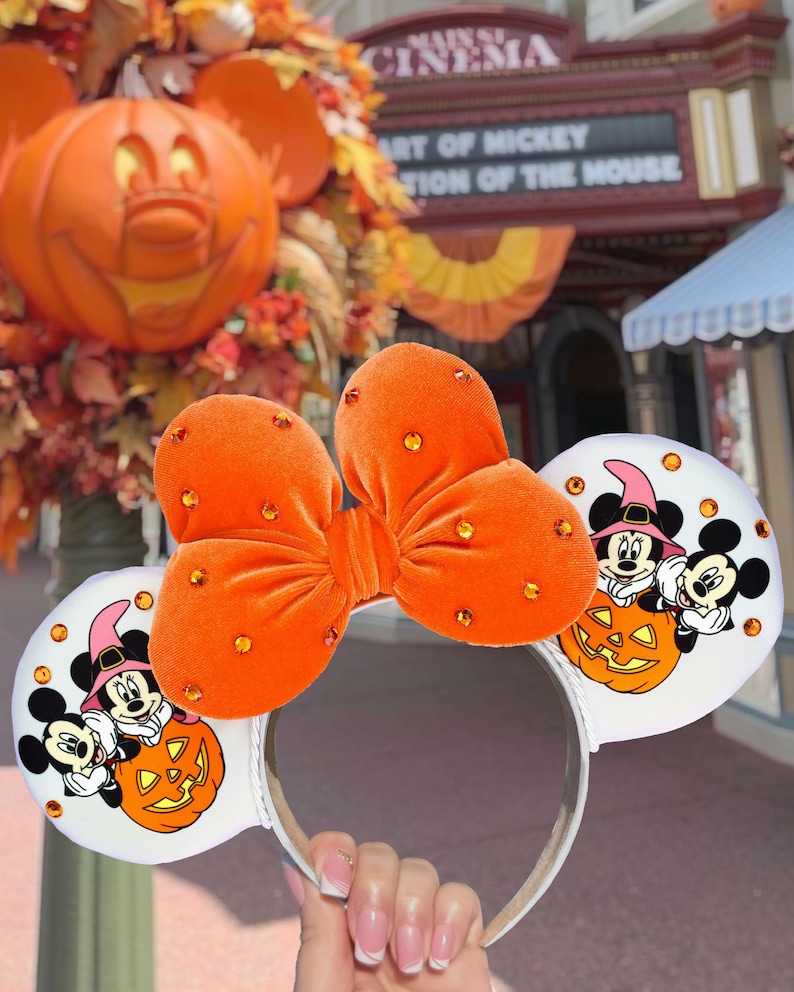 May include: A headband featuring two white ears with a graphic of Mickey and Minnie Mouse in witch hats sitting on a pumpkin. A large orange velvet bow with embedded orange rhinestones sits between the ears. The ears are also accented with orange rhinestones.