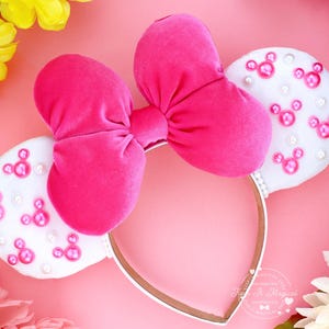 Puede incluir: Una diadema con lazo de terciopelo rosa intenso y orejas de fieltro blanco. Las orejas est&aacute;n decoradas con formas de Mickey Mouse rosas y perlas blancas. La diadema es dorada con ribete blanco. El fondo es rosa con flores.