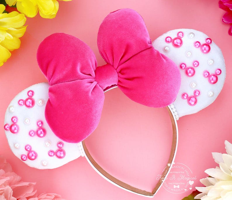 Puede incluir: Una diadema con lazo de terciopelo rosa intenso y orejas de fieltro blanco. Las orejas est&aacute;n decoradas con formas de Mickey Mouse rosas y perlas blancas. La diadema es dorada con ribete blanco. El fondo es rosa con flores.