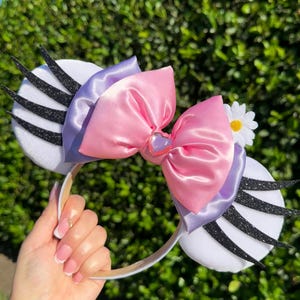 May include: A white headband with large, round, white ears. The ears have black glitter accents. A large pink satin bow is centered on the headband, with a smaller lavender bow underneath. A small white daisy is attached.