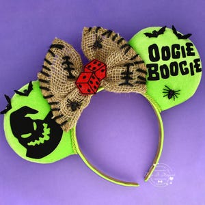 May include: A headband featuring two bright green ears, one with a black silhouette of a character and the other with the text OOGIE BOOGIE. A burlap bow sits in the centre, decorated with black stitching, small spiders, and two red dice.