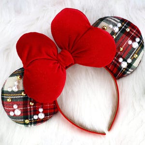 May include: A headband featuring two circular ears covered in a red, green, and white tartan fabric. The ears are embellished with scattered white faux pearls and small gold-toned beads. A large, plush, solid red velvet bow sits between the ears.