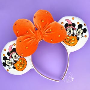 May include: A headband featuring two white circular ears with a graphic of Mickey and Minnie Mouse wearing witch hats behind a jack-o'-lantern. A large orange velvet bow with embedded orange rhinestones sits between the ears.