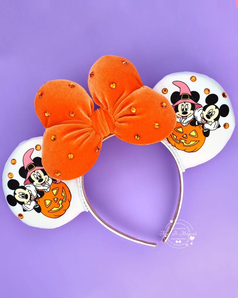 May include: A headband featuring two white circular ears with a graphic of Mickey and Minnie Mouse wearing witch hats behind a jack-o'-lantern. A large orange velvet bow with embedded orange rhinestones sits between the ears.