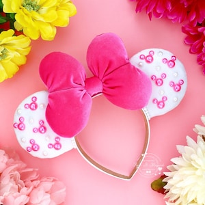 Puede incluir: Una diadema rosa con un lazo grande y dos orejas blancas decoradas con siluetas rosas de Mickey Mouse. La diadema est&aacute; rodeada de flores rosas, amarillas y blancas sobre un fondo rosa.