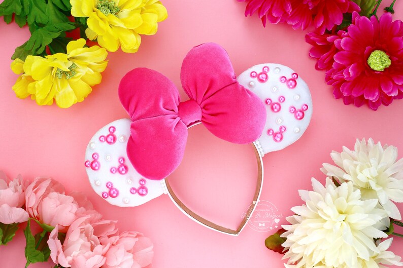 Puede incluir: Una diadema rosa con un lazo grande y dos orejas blancas decoradas con siluetas rosas de Mickey Mouse. La diadema est&aacute; rodeada de flores rosas, amarillas y blancas sobre un fondo rosa.