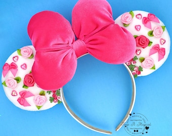 May include: A pink velvet bow headband with white circular ears decorated with pink roses, bows, and hearts. The headband is silver and the background is blue. The ears have the text "Keep It Magical Shop".