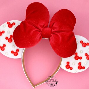 Puede incluir: Diadema con lazo de terciopelo rojo con orejas blancas decoradas con formas de Mickey Mouse rojas y perlas blancas. La diadema tiene una banda dorada. El fondo es rosa.