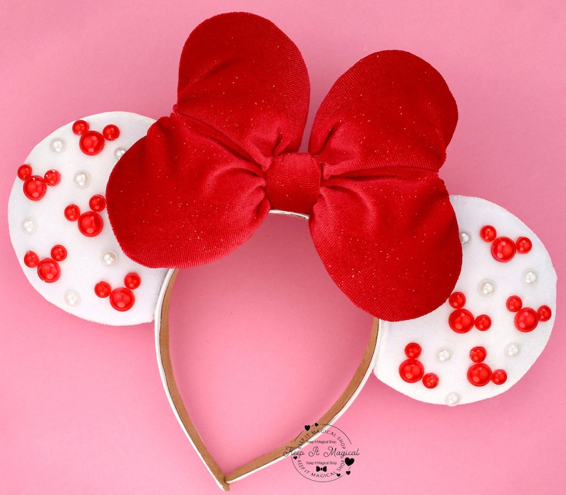 Puede incluir: Diadema con lazo de terciopelo rojo con orejas blancas decoradas con formas de Mickey Mouse rojas y perlas blancas. La diadema tiene una banda dorada. El fondo es rosa.