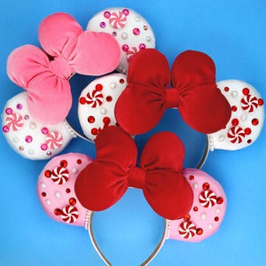May include: A collection of four headbands featuring round ears decorated with white or pink fabric, red and white peppermint candy embellishments, and scattered red, pink, and clear rhinestones. Each headband is topped with a large velvet bow.