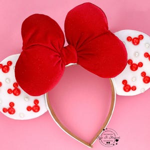 Puede incluir: Una diadema con lazo de terciopelo rojo con orejas blancas decoradas con formas rojas de Mickey Mouse y peque&ntilde;as perlas blancas. La diadema tiene un ribete dorado y un fondo rosa. El texto "Keep It Magical" es visible.