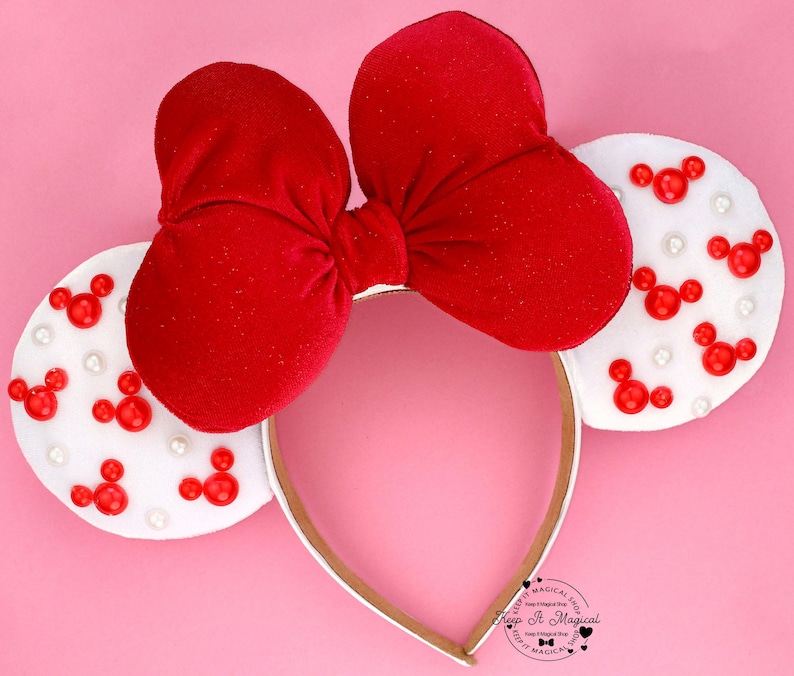 Puede incluir: Una diadema con lazo de terciopelo rojo con orejas blancas decoradas con formas rojas de Mickey Mouse y peque&ntilde;as perlas blancas. La diadema tiene un ribete dorado y un fondo rosa. El texto "Keep It Magical" es visible.