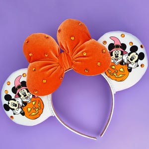 May include: A headband featuring two white circular ears decorated with a graphic of two cartoon mice wearing hats behind a jack-o'-lantern. An orange velvet bow with embedded orange rhinestones sits between the ears on a white headband.