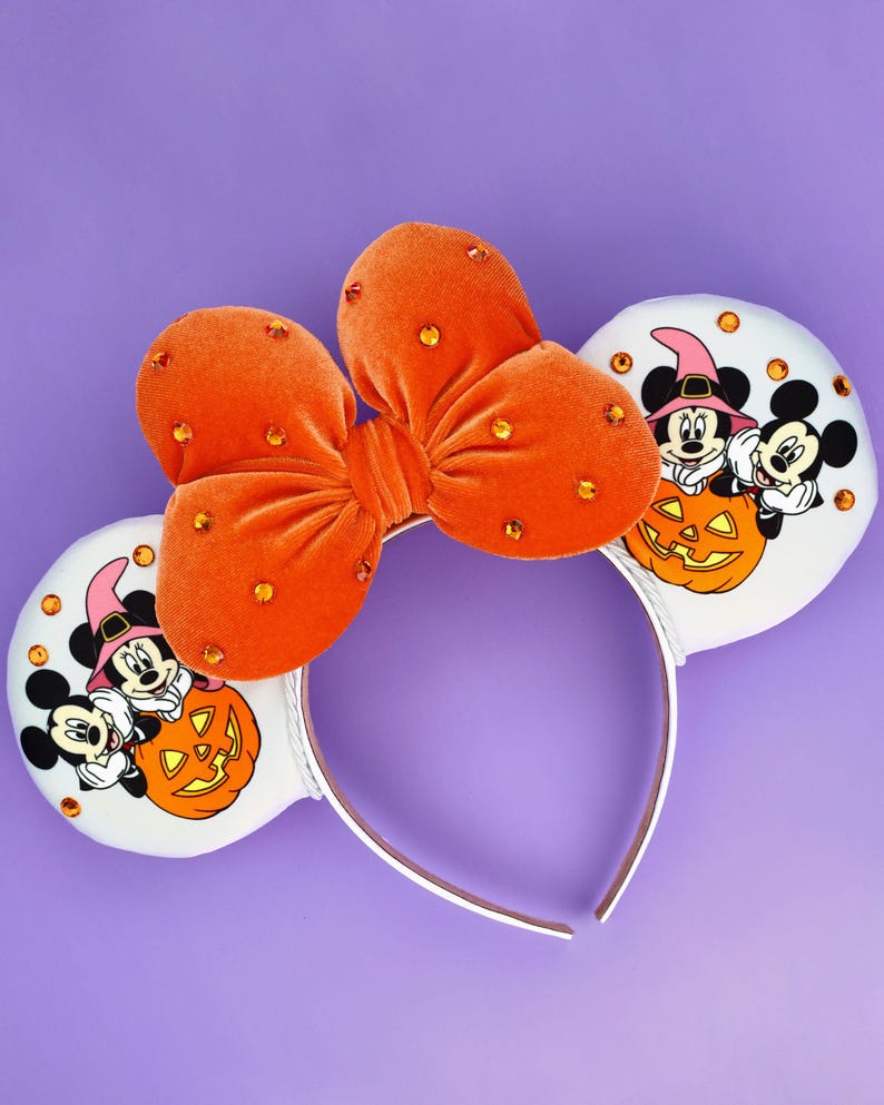May include: A headband featuring two white circular ears decorated with a graphic of two cartoon mice wearing hats behind a jack-o'-lantern. An orange velvet bow with embedded orange rhinestones sits between the ears on a white headband.