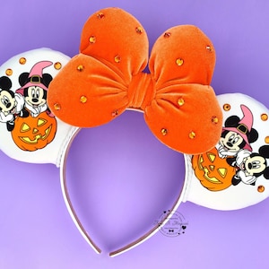 May include: A headband featuring two white ears with illustrations of Mickey and Minnie Mouse wearing witch hats behind a jack-o'-lantern. A large orange velvet bow with orange rhinestones sits between the ears. The text Keep It Magical is visible.
