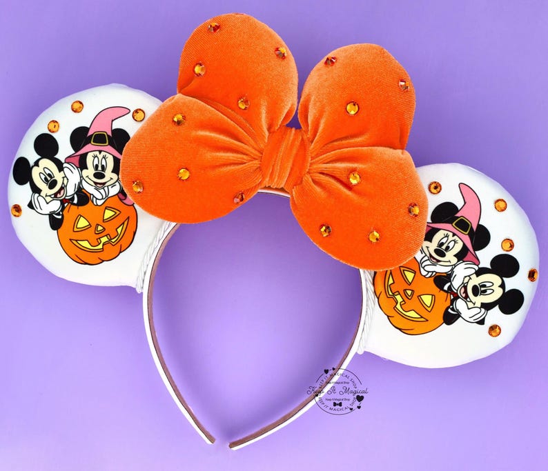 May include: A headband featuring two white ears with illustrations of Mickey and Minnie Mouse wearing witch hats behind a jack-o'-lantern. A large orange velvet bow with orange rhinestones sits between the ears. The text Keep It Magical is visible.
