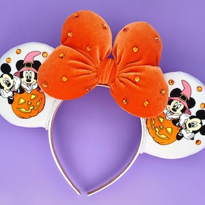May include: A headband featuring two white ears decorated with orange rhinestones and a graphic of Mickey and Minnie Mouse wearing witch hats behind a jack-o'-lantern. A large, plush orange velvet bow with orange rhinestones sits in the centre.