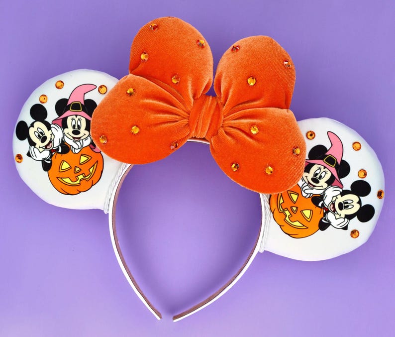 May include: A headband featuring two white ears decorated with orange rhinestones and a graphic of Mickey and Minnie Mouse wearing witch hats behind a jack-o'-lantern. A large, plush orange velvet bow with orange rhinestones sits in the centre.