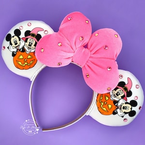 May include: A headband featuring two white ears decorated with a graphic of Mickey and Minnie Mouse wearing witch hats behind an orange jack-o'-lantern. A large, bright pink velvet bow with scattered pink rhinestones sits between the ears.
