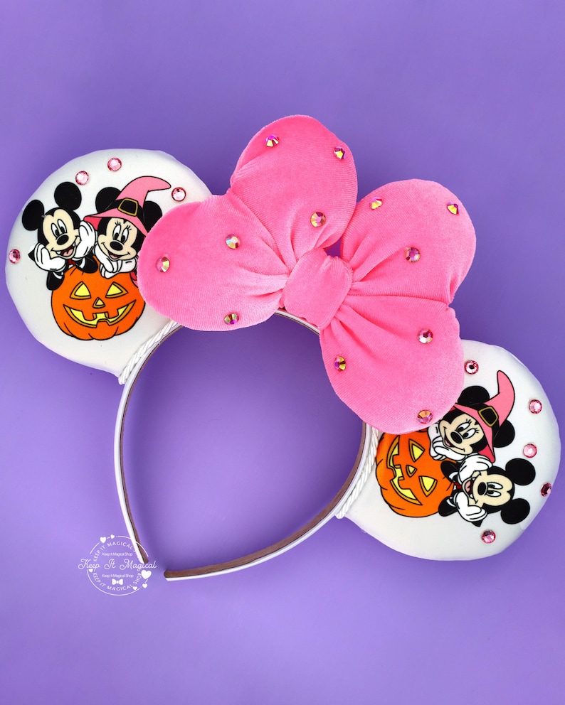 May include: A headband featuring two white ears decorated with a graphic of Mickey and Minnie Mouse wearing witch hats behind an orange jack-o'-lantern. A large, bright pink velvet bow with scattered pink rhinestones sits between the ears.