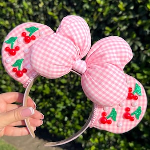 May include: Pink gingham-patterned headband with large bow and mouse ears. The ears are decorated with red cherry and green leaf appliqués, and white pearl accents. The headband is a fun accessory.