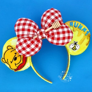 May include: Yellow sequined Minnie Mouse ears with a red and white gingham bow and a wooden honey dipper. One ear features Winnie the Pooh, the other says "HUNNY" with a bee.