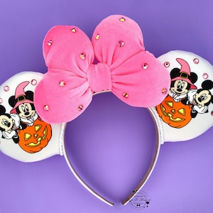 May include: A headband featuring two white circular ears and a large pink velvet bow adorned with scattered pink rhinestones. The ears display a graphic of two cartoon mice wearing witch hats and sitting behind a carved orange jack-o'-lantern.