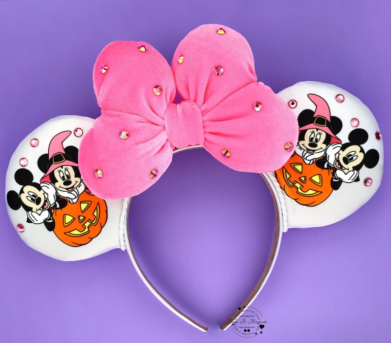 May include: A headband featuring two white circular ears and a large pink velvet bow adorned with scattered pink rhinestones. The ears display a graphic of two cartoon mice wearing witch hats and sitting behind a carved orange jack-o'-lantern.