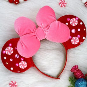 May include: A headband featuring two round red velvet ears and a large central pink velvet bow. The ears are decorated with white and pink peppermint candy embellishments, small pink pearls, and clear faceted rhinestones.