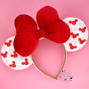Puede incluir: Una diadema con lazo de terciopelo rojo y orejas de fieltro blanco. Las orejas est&aacute;n decoradas con formas rojas de Mickey Mouse y peque&ntilde;as perlas blancas. La diadema es dorada y est&aacute; sobre un fondo rosa.