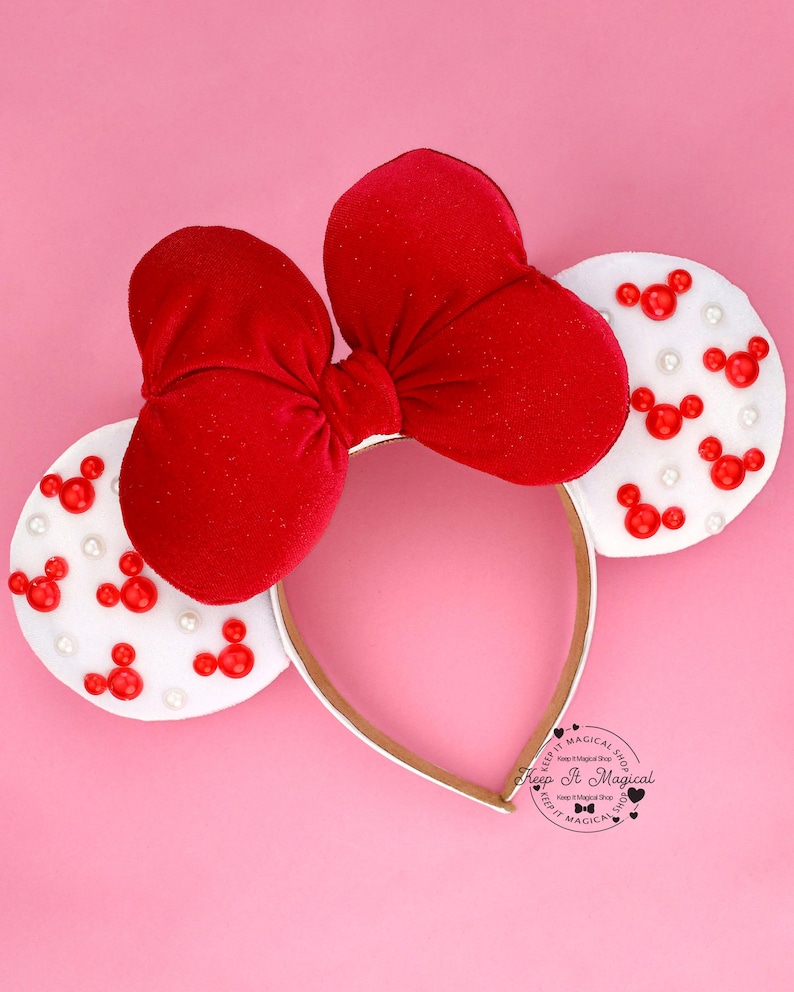 Puede incluir: Una diadema con lazo de terciopelo rojo y orejas de fieltro blanco. Las orejas est&aacute;n decoradas con formas rojas de Mickey Mouse y peque&ntilde;as perlas blancas. La diadema es dorada y est&aacute; sobre un fondo rosa.
