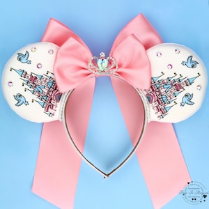 May include: A headband featuring two white circular ears decorated with illustrations of a pink castle and blue birds, accented with small crystals. A large pink satin bow sits between the ears, topped with a small silver-toned crown charm.