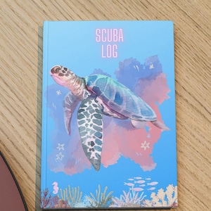 Puede incluir: Una libreta azul con una ilustración de acuarela de una tortuga marina nadando en un cielo rosa y azul. La libreta dice "SCUBA LOG" en la portada.