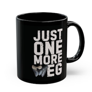 May include: Black ceramic coffee mug with white text that reads "Just One More" and a graphic of a shark tooth.