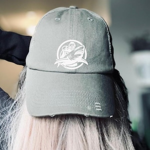 May include: A green baseball cap featuring a white graphic of a shark and a scuba diver. The cap has a distressed look and is made of a soft, comfortable fabric.