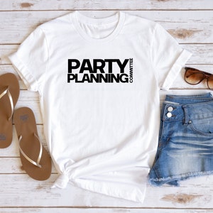 May include: White t-shirt with the text "PARTY PLANNING COMMITTEE" in black. The shirt is paired with denim shorts, brown sandals, and sunglasses. The items are arranged on a light wood-grain surface.