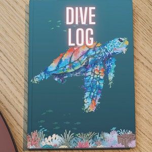Op de afbeelding: Tealblauwe omslag met de tekst "DIVE LOG" in roze. Een kleurrijk aquarel schilderij van een zeeschildpad die in de oceaan zwemt met koraal en vissen op de bodem.