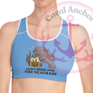 May include: A light blue sports bra with a white trim and a cartoon manatee graphic. The manatee is gray and purple, leaning on brown seaweed. Text on the bra reads "JUST HERE FOR THE SEAGRASS."