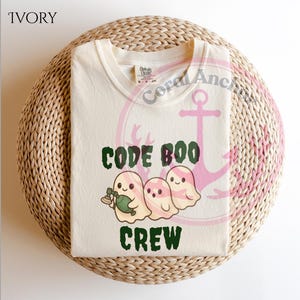 May include: Ivory-colored t-shirt with the words "CODE BOO CREW" in green and a graphic of three cartoon ghosts. One ghost holds a green bottle. The shirt is folded on a woven, circular surface.